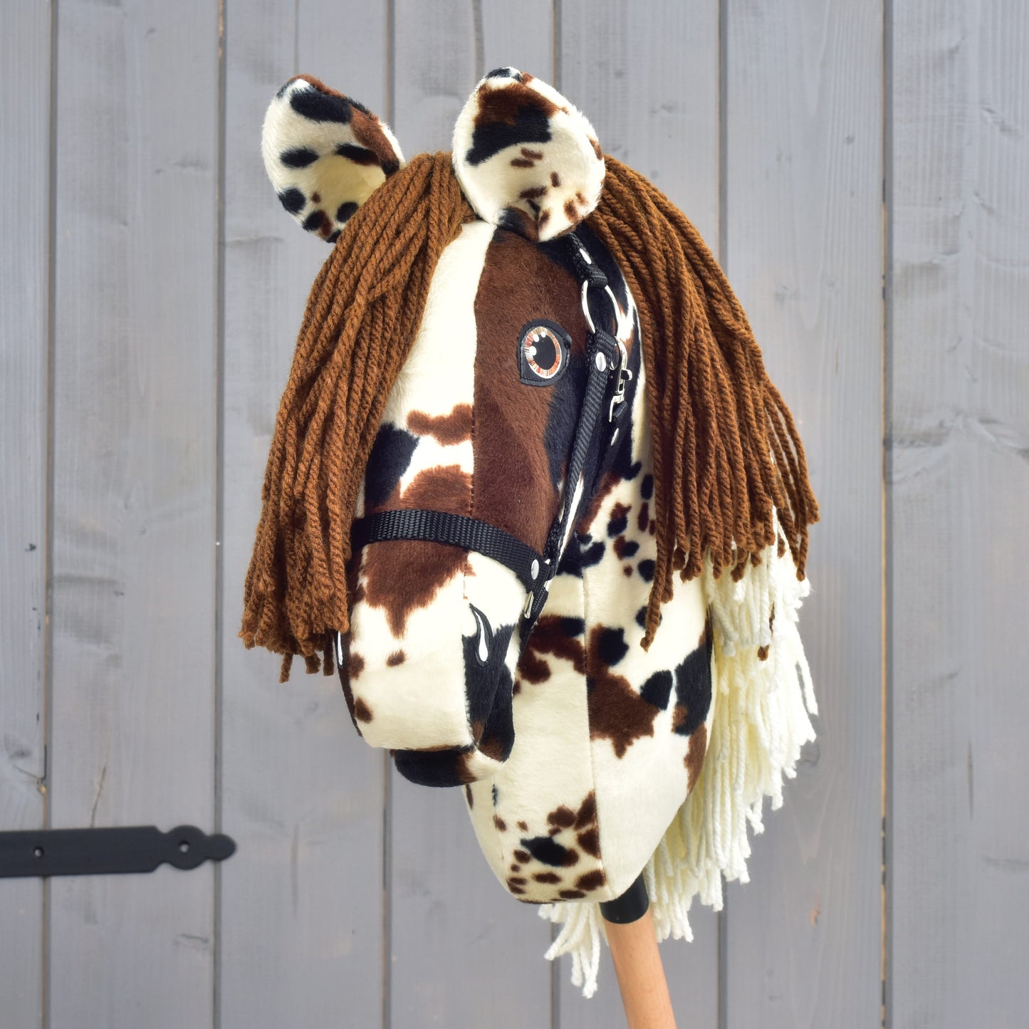 The Hobby Horse Dakota with Black Halter by Hobby Horse LarDen is a handcrafted hobby horse on a stick, inspired by the wild spirit of the Mustang. It features a brown and white spotted plush head with brown yarn bangs and a white yarn mane, wears a black halter, and is set against rustic gray wooden planks.