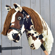 A plush Hobby Horse LarDen "Dakota with Black Halter," inspired by the mustang with a brown yarn mane, black halter, and a spotted brown-and-white coat that captures the wild spirit of North America—beautifully set against a wooden background.