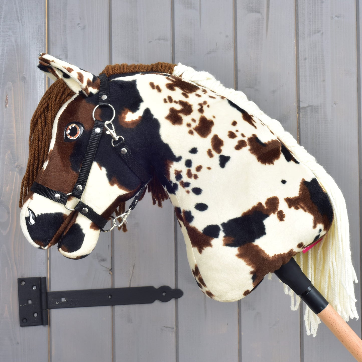 The Hobby Horse Dakota with Black Halter by Hobby Horse LarDen—a plush stick horse featuring brown and white spots, a bridle, and a yarn mane inspired by the wild Mustang horses of North America—is displayed against a gray wooden wall background.