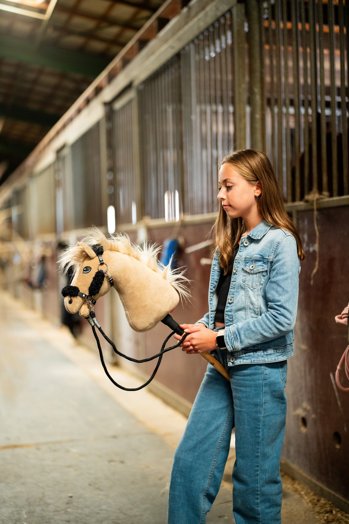 Dziewczynka w dżinsowej kurtce prowadzi Hobby Horse Dancing Lady z czarnym kantarem od Hobby Horse LarDen stajennym korytarzem – kremowa pluszowa głowa, jasna grzywa i grzywka z włóczki – pokazany z czarnym kantarem ze skóry ekologicznej z pełnym czarnym futerkiem i czarnymi wodzami