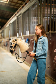 Dziewczynka w dżinsowej kurtce prowadzi Hobby Horse Dancing Lady z czarnym kantarem od Hobby Horse LarDen stajennym korytarzem – kremowa pluszowa głowa, jasna grzywa i grzywka z włóczki – pokazany z czarnym kantarem ze skóry ekologicznej z pełnym czarnym futerkiem i czarnymi wodzami