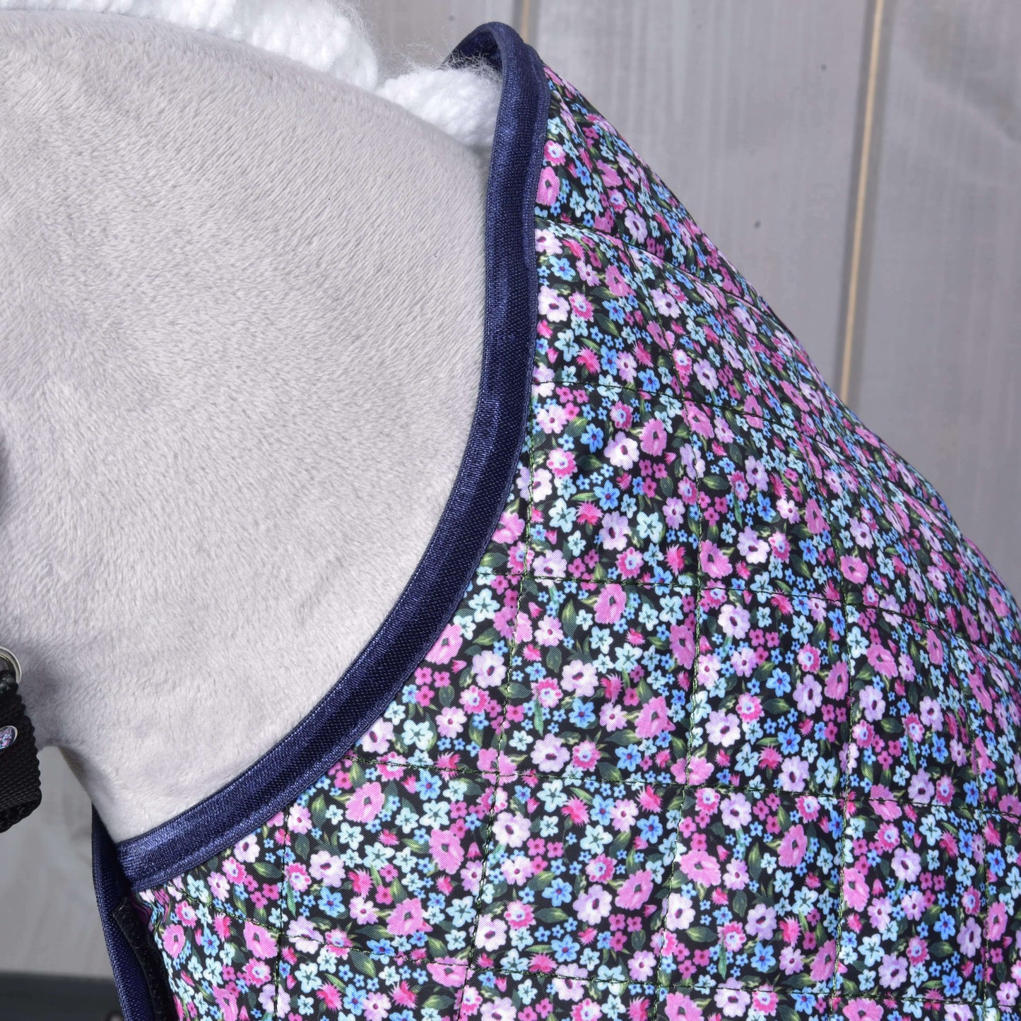 Zbliżenie na konia noszącego pikowaną derkę Flower od Hobby Horse LarDen z granatowym obszyciem i drobnymi różowymi, fioletowymi, białymi oraz niebieskimi kwiatami na ciemnym tle. Tło stanowi jasne drewno.