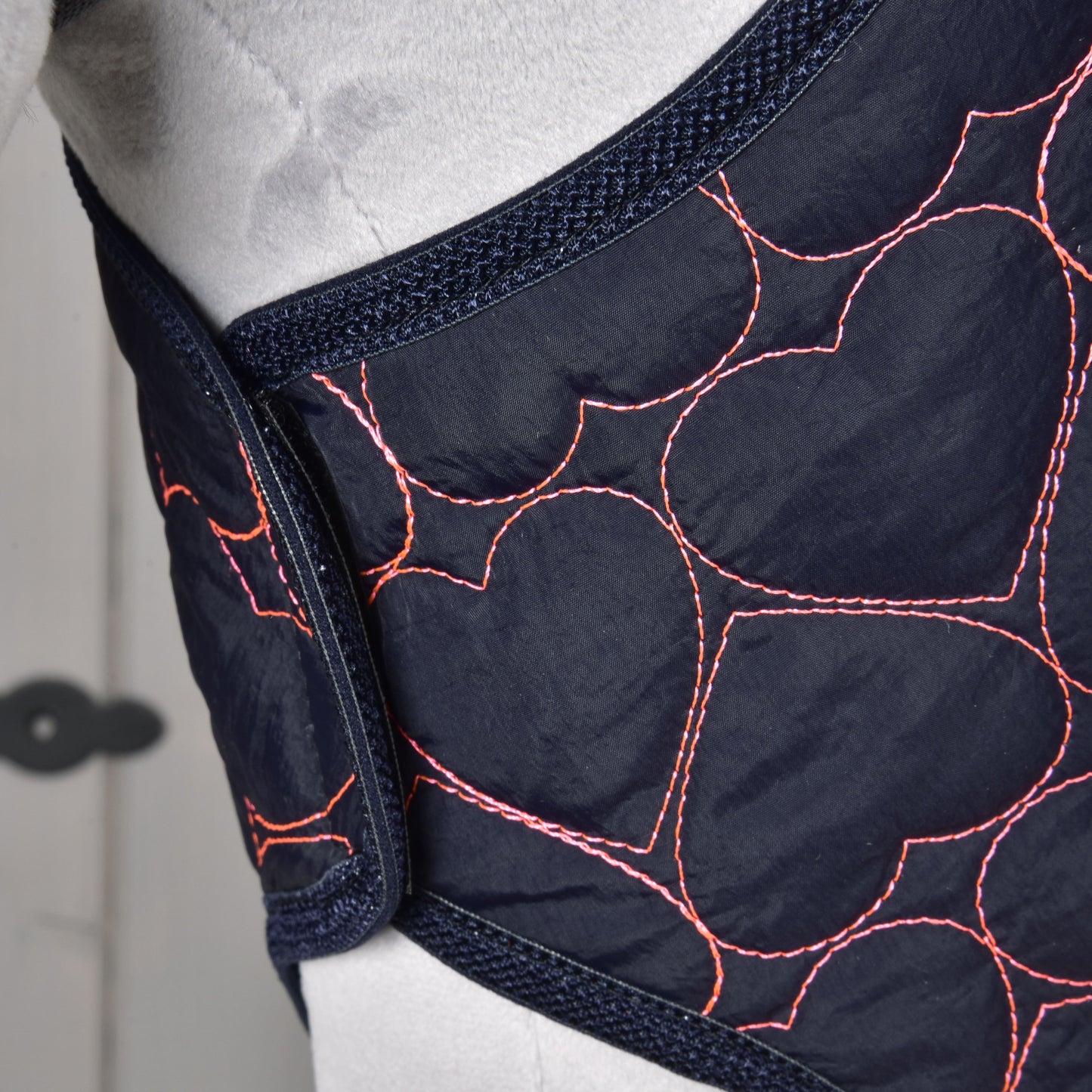 Close-up of the Hobby Horse LarDen “Blanket Pink Heart,” featuring dark blue quilted fabric with pink heart-shaped stitching, wrapped around a light gray surface, suggesting it is part of a warm hobby horse blanket.