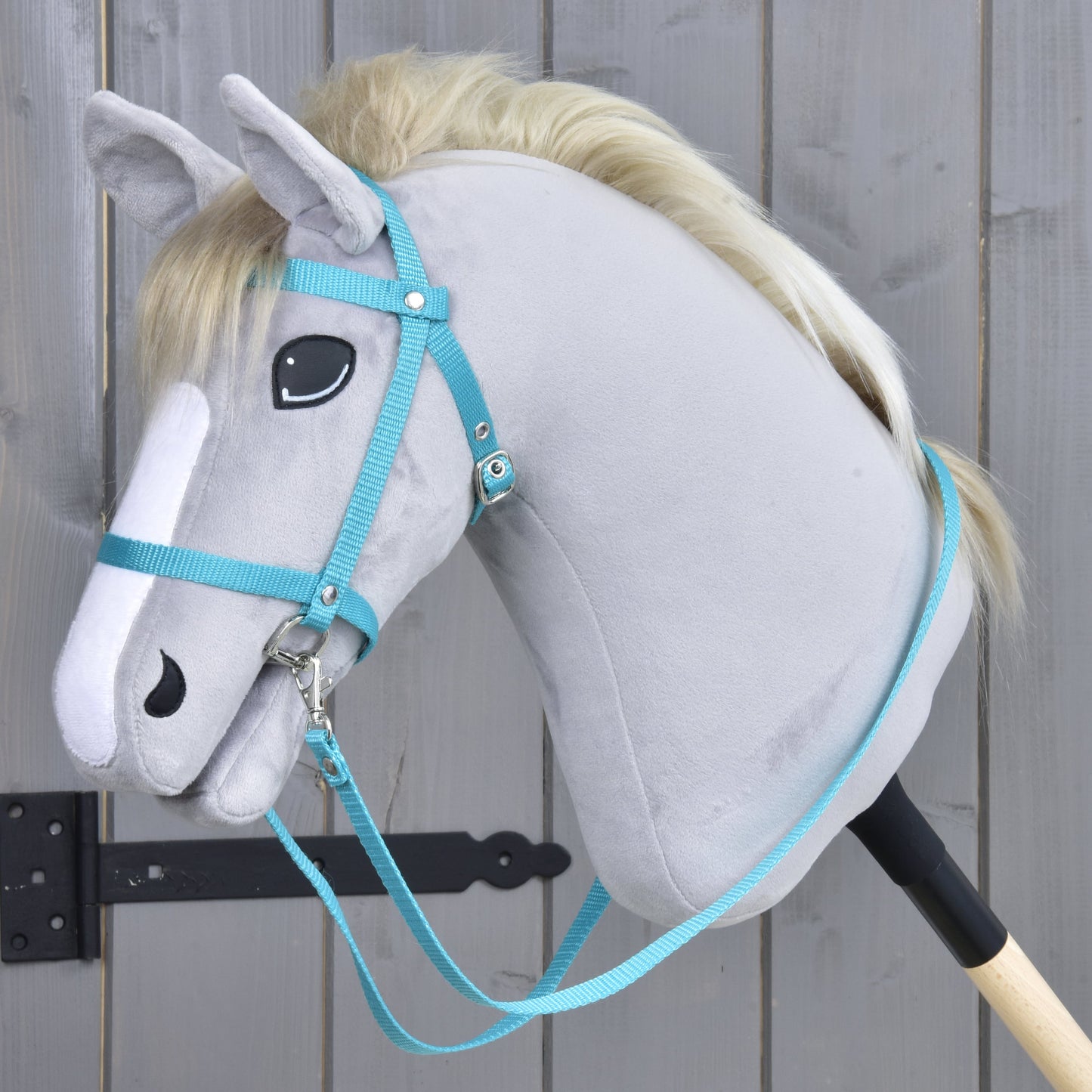 A gray plush hobby horse with a light mane, wearing the Hobby Horse LarDen black bridle with reins from strap turquoise, is mounted on a wooden stick and placed before a gray wooden fence.