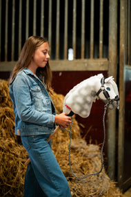 Dziewczynka w dżinsowej kurtce stoi przed belami słomy w stajni, trzymając Hobby Horse Elektra z czarnym kantarem od Hobby Horse LarDen – biała dereszowata pluszowa głowa, niebieskie oko, biała grzywa z włóczki – pokazany z kantarem Graphite Full z futerkiem ze skóry ekologicznej i dzierganym uwiązem