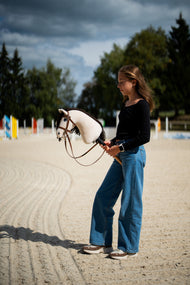 Dziewczynka w czarnej bluzce stoi na piaszczystym placu pod burzowym niebem, trzymając Hobby Horse Frederik LD z czarnym kantarem od Hobby Horse LarDen, z kolorowymi drągami do skoków w tle – kremowa pluszowa głowa, ciemnobrązowa grzywa z włóczki – pokazany z kompletnym brązowym ogłowiem z nachrapnikiem