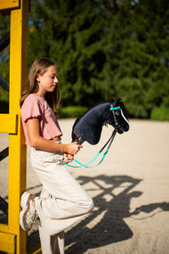 Dziewczynka w różowej bluzce opiera się o żółty stojak do skoków, trzymając Hobby Horse Freya z czarnym kantarem od Hobby Horse LarDen – czarna pluszowa głowa, biała łysina, czarna grzywa z włóczki – pokazany z kompletnym turkusowym ogłowiem bez nachrapnika
