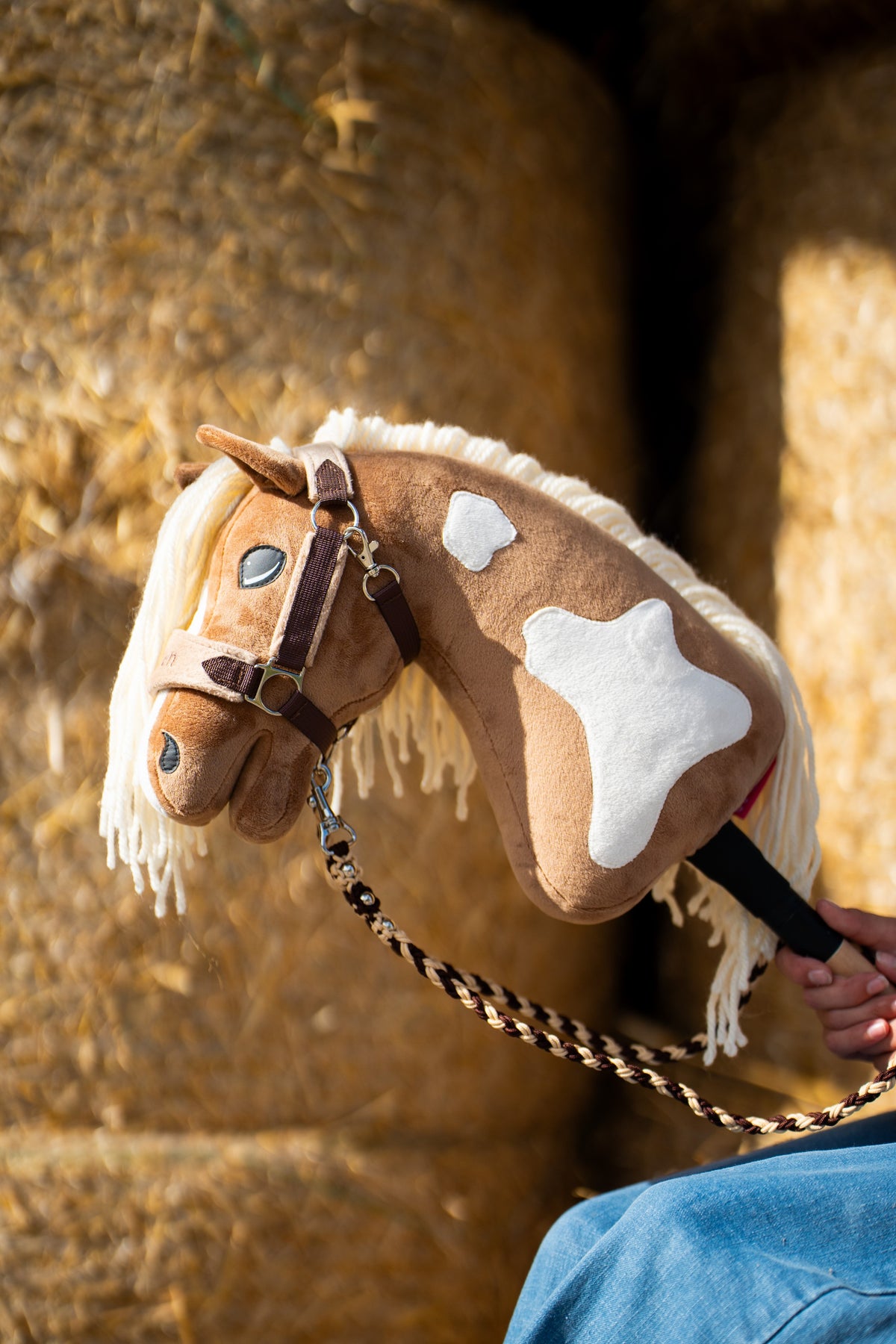 Głowa i szyja Hobby Horse Fury z czarnym kantarem od Hobby Horse LarDen przed okrągłymi belami słomy – palomino plusz z białymi znaczeniami, kremowa grzywa z włóczki – pokazany z podszytym futerkiem kantarem LarDen Brown i plecionym uwiązem Bloom Brown