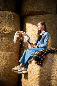Dziewczynka w dżinsowej kurtce siedzi wysoko na okrągłych belach słomy, trzymając Hobby Horse Fury z czarnym kantarem od Hobby Horse LarDen – palomino pluszowa głowa z białymi znaczeniami, kremowa grzywa z włóczki – pokazany z podszytym futerkiem kantarem LarDen Brown i uwiązem Bloom Brown