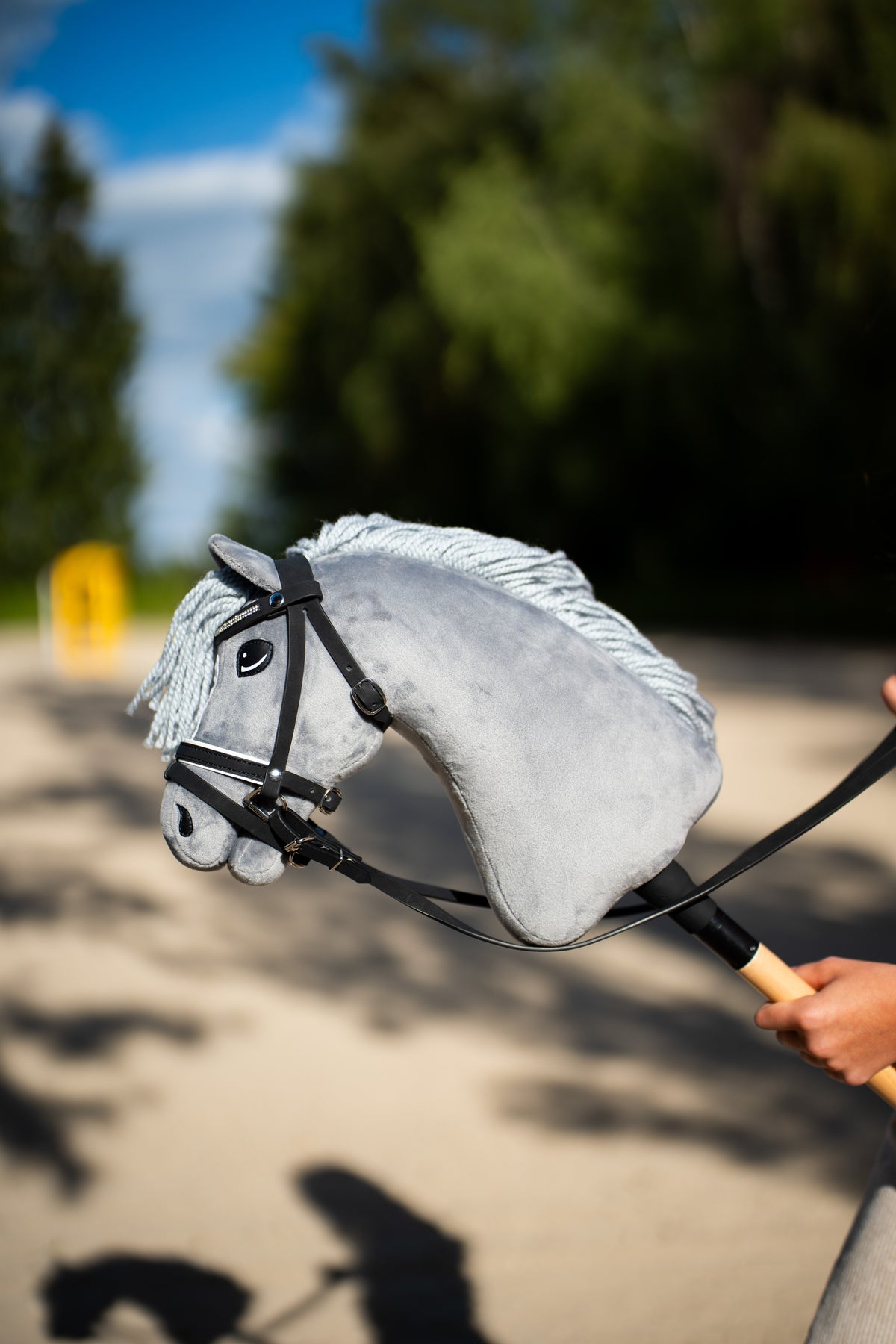 Detail photo: the head of the Hobby Horse Grace with Black Halter by Hobby Horse LarDen held up on a sand arena against dark conifers - grey plush, pale grey yarn mane and forelock - shown here with the complete De Luxe bridle with two browbands.