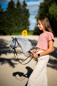 Dziewczynka w różowej bluzce prowadzi Hobby Horse Grace z czarnym kantarem od Hobby Horse LarDen przez piaszczysty plac – szara pluszowa głowa, jasnoszara grzywa z włóczki – pokazany z kompletnym ogłowiem De Luxe z dwoma naczółkami