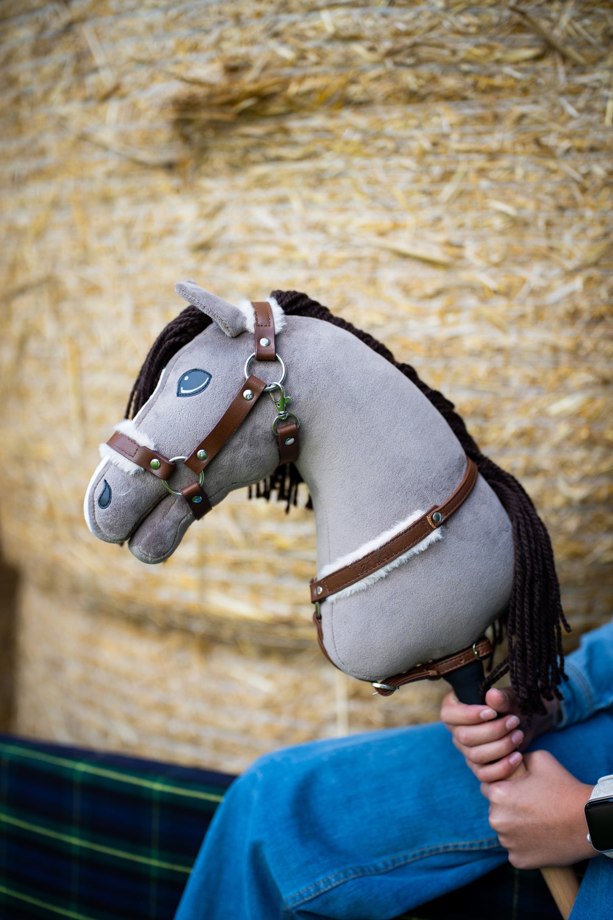 Detail photo: the head of the Hobby Horse Guccini by Hobby Horse LarDen in front of straw bales - grey plush, white nose stripe, dark brown yarn mane - shown here with the fur-lined Walnut halter and the Walnut breastgirth with fur.