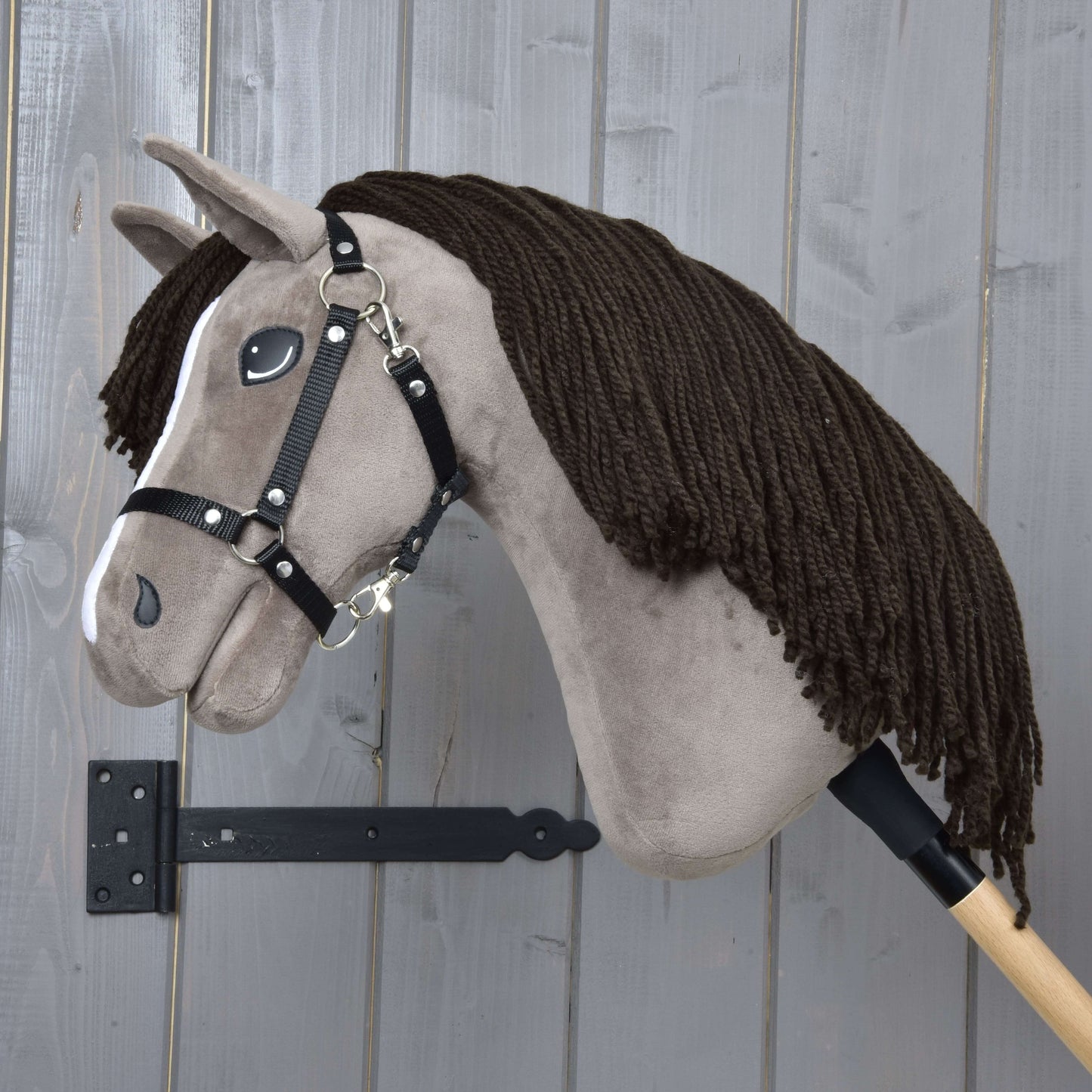 The Hobby Horse Guccini with Black Halter by Hobby Horse LarDen features a brown yarn mane and wooden stick, set against a gray wall—ideal for young equestrian sport fans.