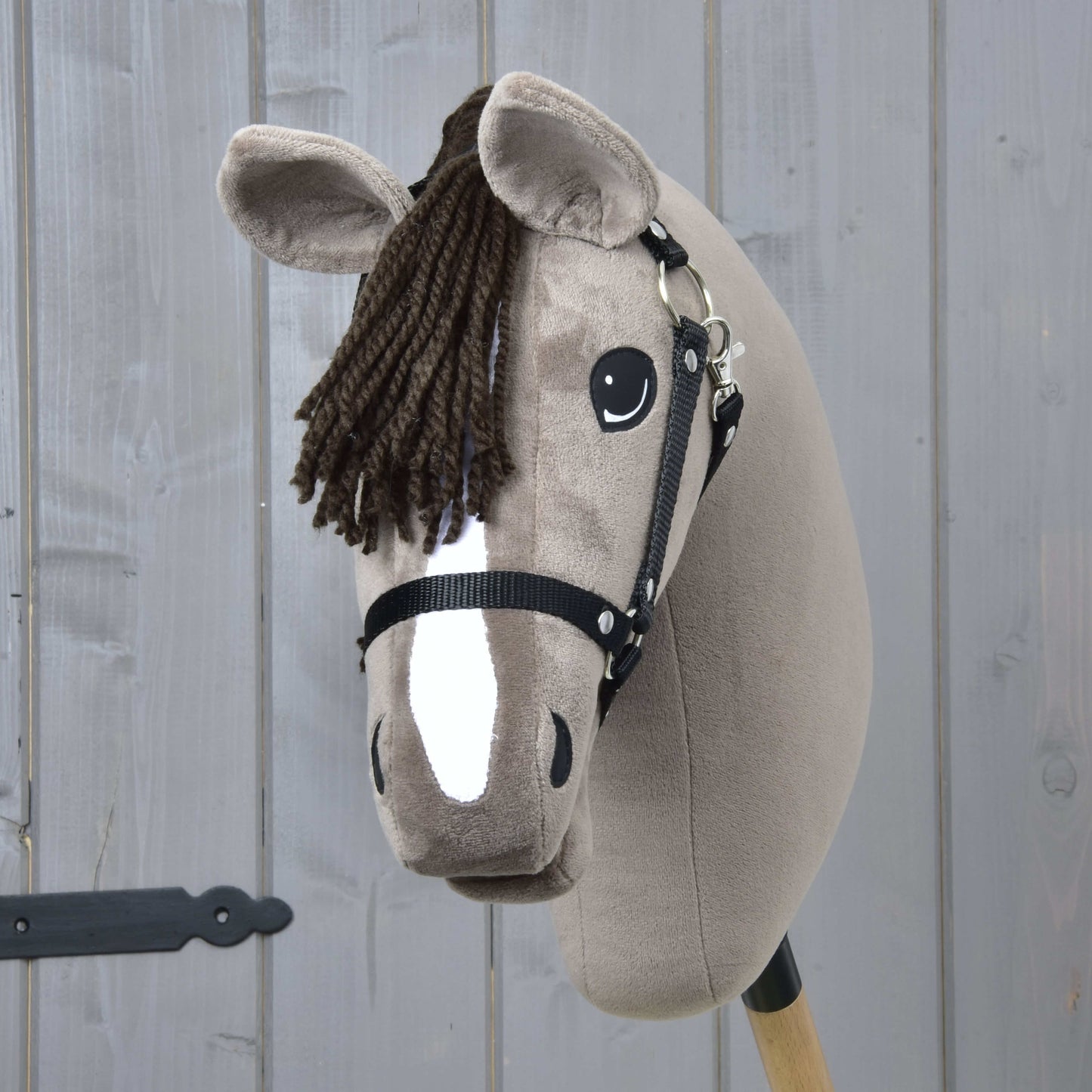 The Hobby Horse LarDen Guccini with Black Halter features a brown yarn mane and white blaze, mounted on a wooden stick—perfect for equestrian play and proudly displayed against a gray wood wall.