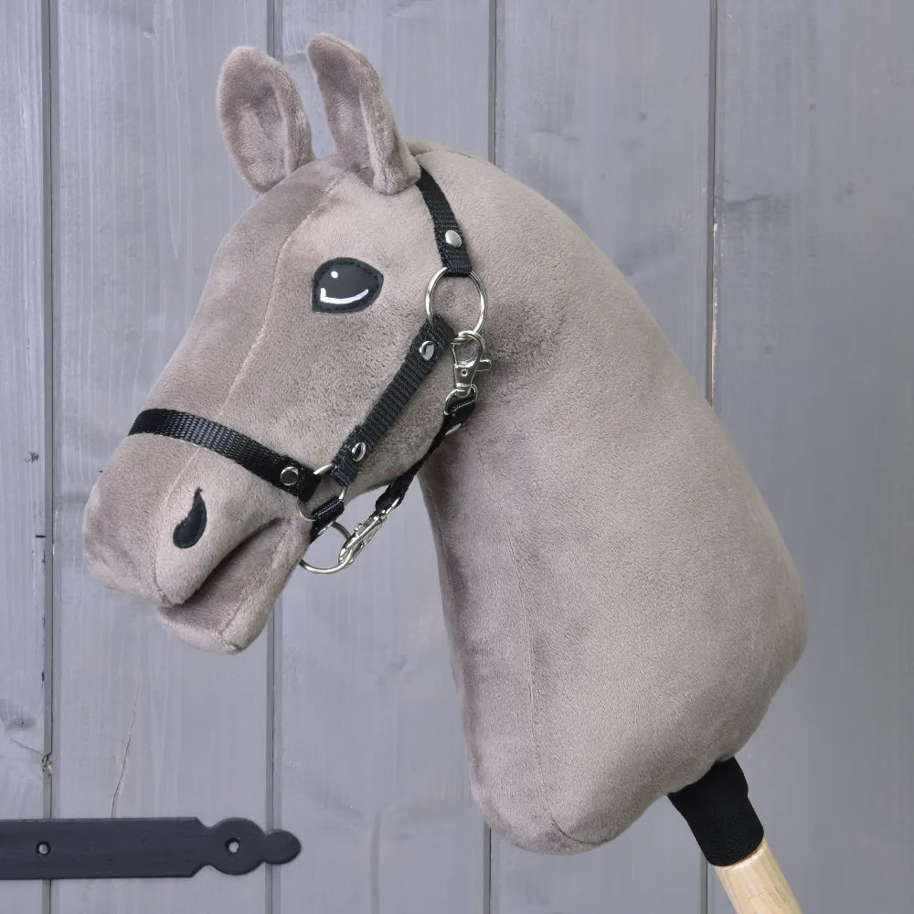 Zbuduj swojego konia od related_to_12441055625556, personalizowany Hobby Horse z szarą pluszową głową, wyrazistymi niebieskimi oczami i czarną uzdą, osadzony na drewnianym kiju, pokazany na tle szarych drewnianych drzwi.