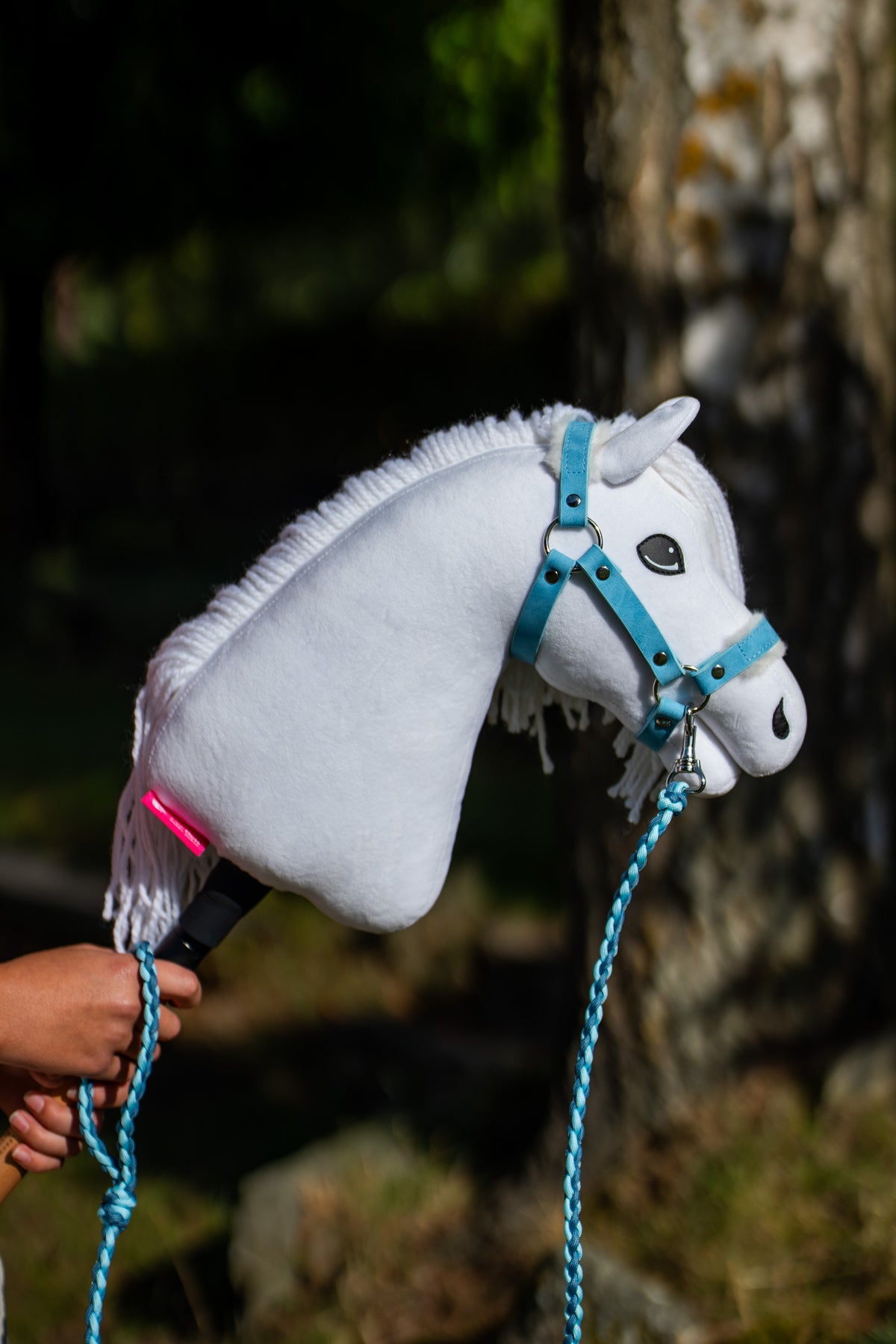 Detail photo: the head of the Hobby Horse Lady with Black Halter by Hobby Horse LarDen held up in dappled shade - white plush, dark eye, white yarn mane - shown here with the Pastell Blue fur halter in leatherette and the Double Blue knitted lead rope.