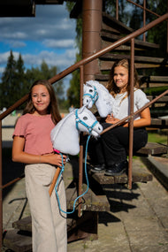 Dwie dziewczyny z białymi Hobby Horses od Hobby Horse LarDen stoją na zardzewiałych metalowych schodach, a z przodu jest Hobby Horse Lady z kantarem Black Halter – biała pluszowa głowa, biała grzywa z włóczki – pokazany z kantarem z futerkiem Pastell Blue ze skóry ekologicznej i plecionym Uwiązem Double Blue