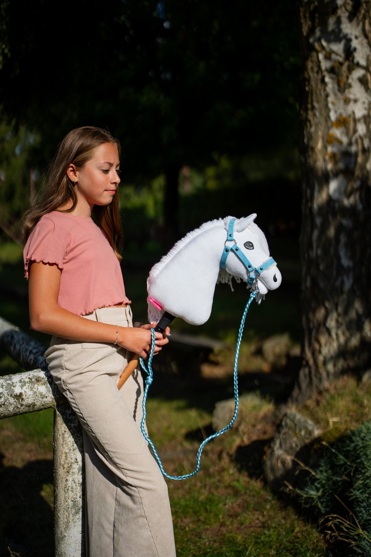 Lifestyle photo: a girl in a pink top stands by a mossy tree holding the Hobby Horse Lady with Black Halter by Hobby Horse LarDen, the lead rope hanging in a loop - white plush head, white yarn mane - shown here with the Pastell Blue fur halter in leatherette and the Double Blue knitted lead rope.