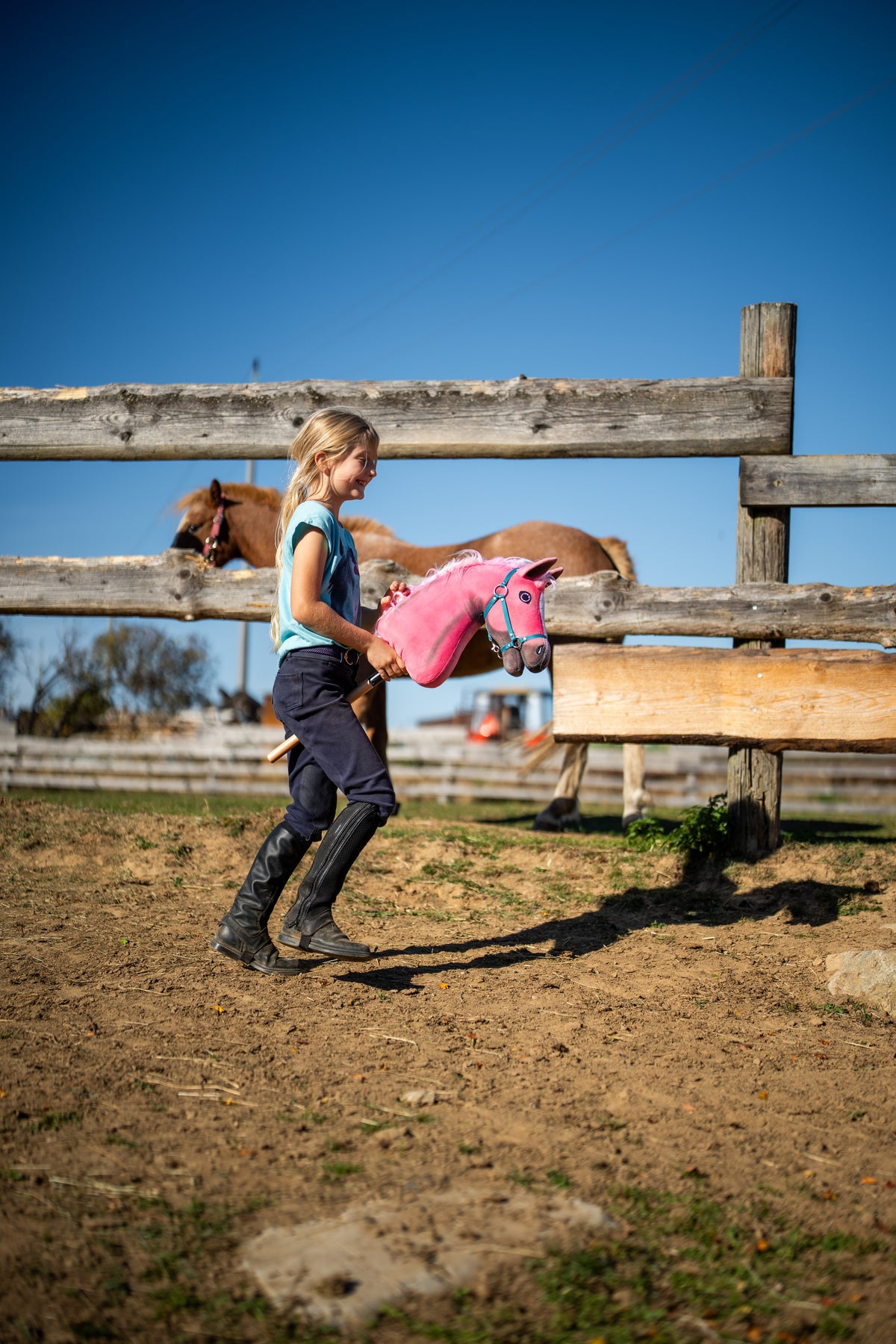Uśmiechnięta dziewczynka w oficerkach prowadzi po padoku Hobby Horse Pinkie, tu z turkusowym kantarem, obok kasztanowatego konia