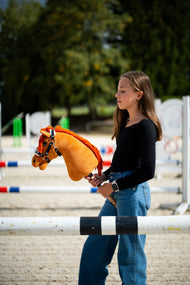 Dziewczynka w czarnej bluzce stoi za drągiem do skoków na piaszczystym placu, trzymając Hobby Horse Little Angel z czarnym kantarem od Hobby Horse LarDen – kasztanowata pluszowa głowa, jaskrawoczerwona grzywa z włóczki – pokazany z kantarem Espresso z futerkiem ze skóry ekologicznej