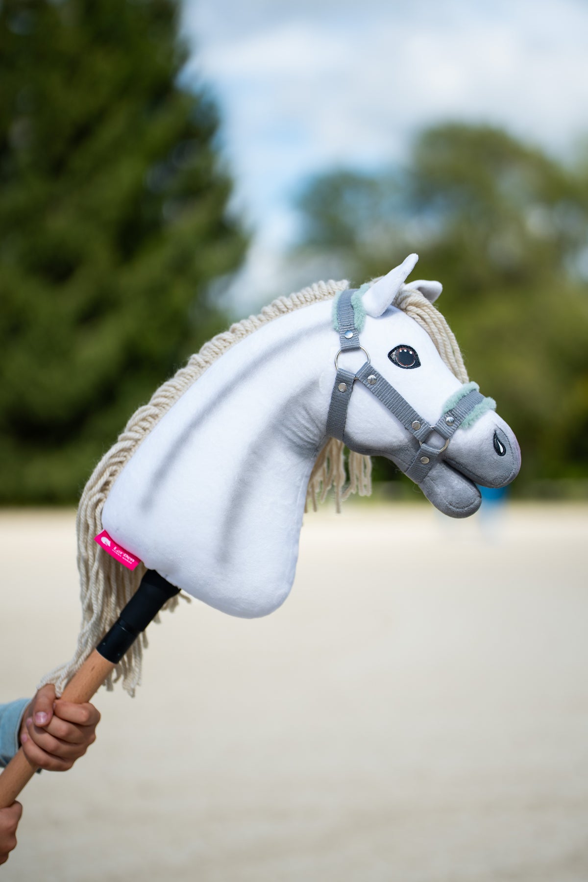 Detail photo: the head of the Hobby Horse Little Lady with Black Halter by Hobby Horse LarDen held up on a sand arena, conifers behind - white plush, cream yarn mane and forelock - shown here with the grey halter with mint fur.
