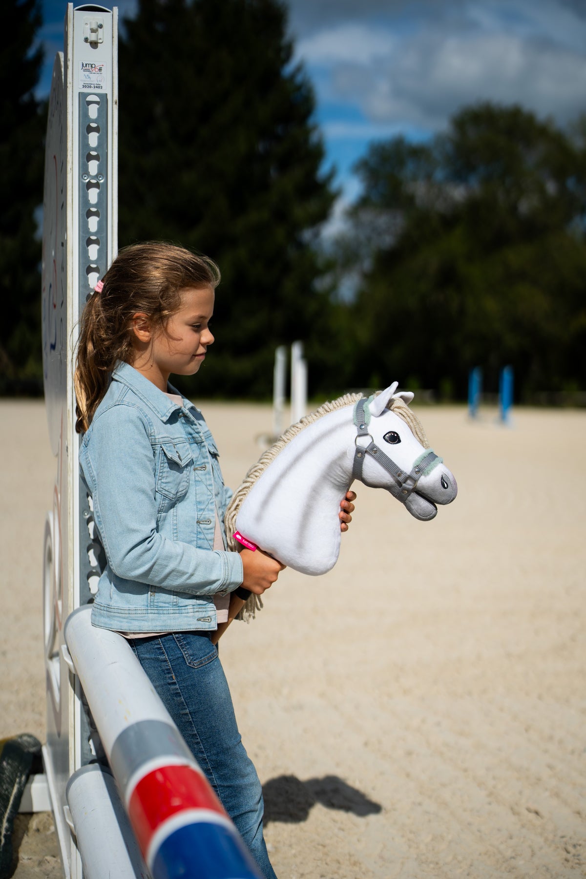 Lifestyle photo: a girl in a denim jacket stands by a jump wing on a sand arena holding the Hobby Horse Little Lady with Black Halter by Hobby Horse LarDen - white plush head, cream yarn mane - shown here with the grey halter with mint fur.