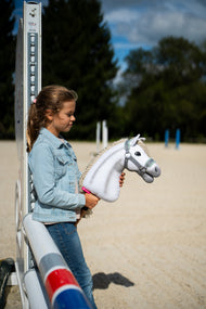 Dziewczynka w dżinsowej kurtce stoi przy stojaku do skoków na piaszczystym placu, trzymając Hobby Horse Little Lady z czarnym kantarem od Hobby Horse LarDen – biała pluszowa głowa, kremowa grzywa z włóczki – pokazany z szarym kantarem z miętowym futerkiem