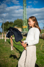 Dziewczyna w białym swetrze stoi na nasłonecznionej łące, trzymając Hobby Horse Loki z kantarem Black Halter od Hobby Horse LarDen – czarna pluszowa głowa, biała łysina, czarna grzywa z włóczki – pokazany z kompletnym Ogłowiem Pure Black bez Nachrapnika