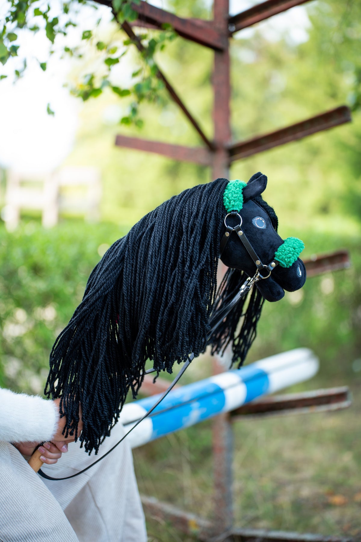 Głowa i długa grzywa Hobby Horse Maximus z kantarem Black Halter od Hobby Horse LarDen, uniesione przy drewnianej przeszkodzie, z zielenią w tle – czarny plusz, czarna grzywa z włóczki – pokazany z czarnym kantarem ze skóry ekologicznej z zielonym futerkiem i czarnymi Wodzami ze skóry ekologicznej
