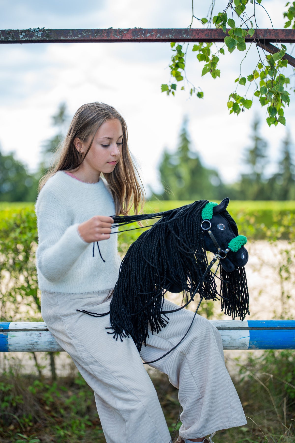 Dziewczyna w jasnym swetrze siedzi na ogrodzeniu padoku, zbierając Wodze Hobby Horse Maximus z kantarem Black Halter od Hobby Horse LarDen – czarna pluszowa głowa, długa czarna grzywa z włóczki – pokazany z czarnym kantarem ze skóry ekologicznej z zielonym futerkiem i czarnymi Wodzami ze skóry ekologicznej