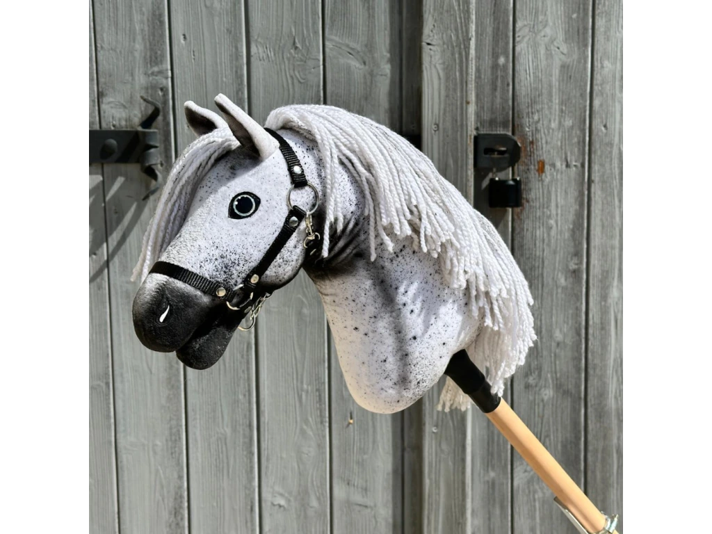 Hobby Horse Naivy z czarnym kantarem - rozmiar S od Hobby Horse LarDen z ręcznie wykonaną biało-szarą cętkowaną głową, białą grzywą z włóczki i czarnym kantarem na drewnianym kiju, pokazany na tle postarzałych szarych drewnianych drzwi.