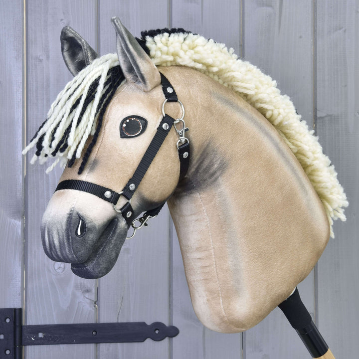 Zbliżenie na beżowego Hobby Horse Ozzy z czarnym kantarem od Hobby Horse LarDen z czarno-białą włóczkową grzywą, czarnym kantarem i guzikowymi oczami, osadzonego na kiju na tle szarego drewna.