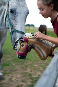 Prawdziwy szary koń opuszcza głowę, by spotkać Hobby Horse Ozzy z kantarem Black Halter od Hobby Horse LarDen, trzymanego przez dziewczynę w kasztanowej bluzce – gniada pluszowa głowa z ciemnym cieniowaniem, kremowo-czarna grzywa z włóczki – pokazany z podszytym futerkiem kantarem Teddy Maroon