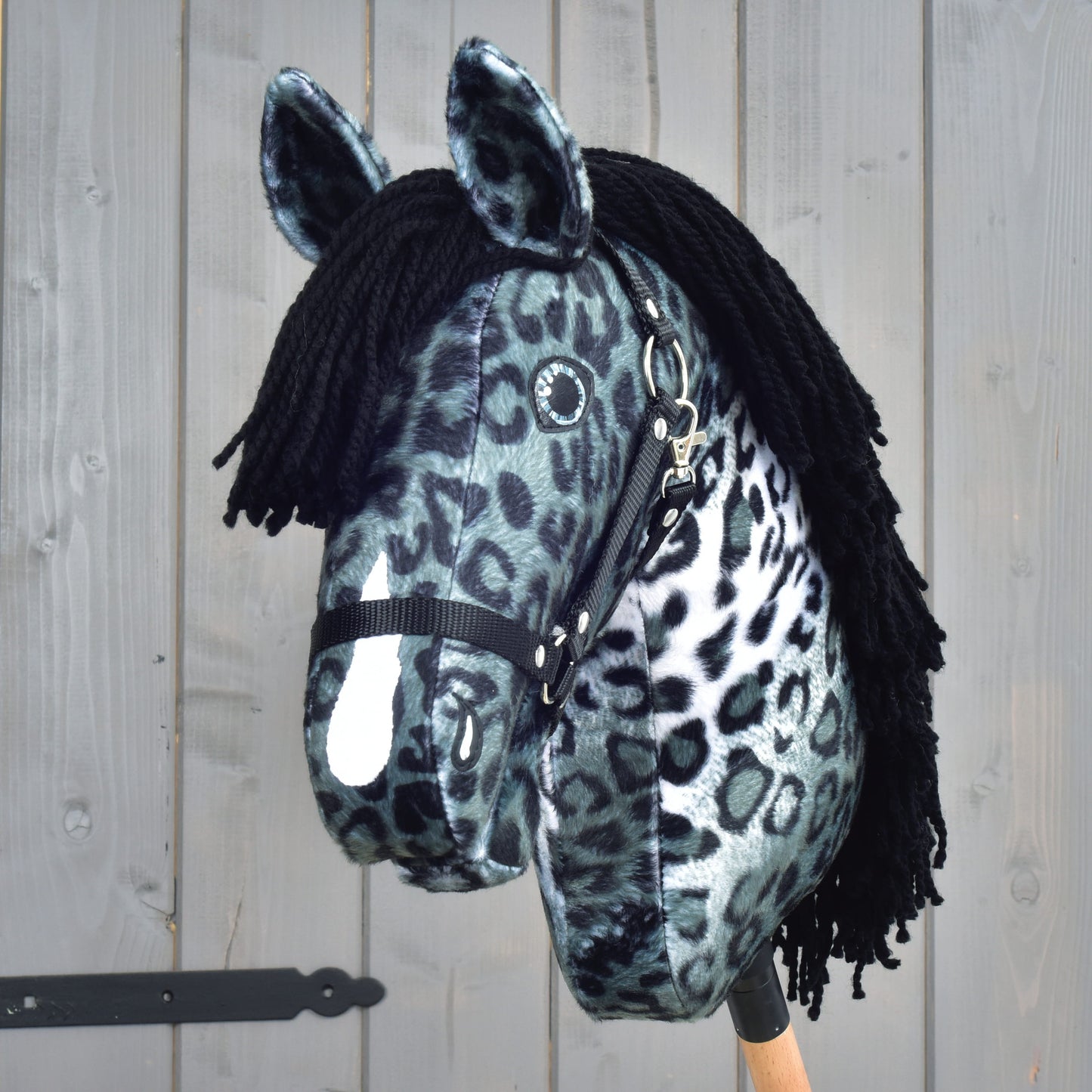 The Hobby Horse LarDen "Hobby Horse Picasso with Black Halter" is a plush hobby horse designed to resemble an Appaloosa stallion, showcasing a bold black and white leopard-spot pattern that echoes the look of a spotted coat. It features a black yarn mane, a black halter, and comes mounted on a wooden stick, pictured against a wooden panel background.