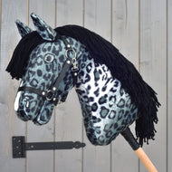 Hobby Horse LarDen "Picasso" z czarnym kantarem, stylizowany na efektownego ogiera, z czarno-białą tkaniną w cętkowany wzór lamparta przypominający umaszczenie appaloosy, z czarną włóczkową grzywą i czarnym kantarem, osadzony na drewnianym kiju na tle szarych drewnianych drzwi.