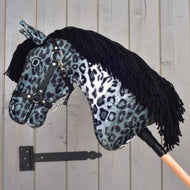 Hobby Horse Picasso z czarnym kantarem od Hobby Horse LarDen, z pluszową głową w szaro-czarne cętki inspirowane umaszczeniem appaloosy, z czarnym kantarem i czarną włóczkową grzywą, osadzony na drewnianym kiju na tle szarej drewnianej ściany.