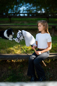 Dziewczyna w białej bluzce siedzi na ławce w ogrodzie w wieczornym słońcu, trzymając Hobby Horse Popcorn z kantarem Black Halter od Hobby Horse LarDen, a za nią odpoczywa border collie – biała pluszowa głowa, biała grzywa z włóczki – pokazany z ozdobnym Ogłowiem bezwędzidłowym