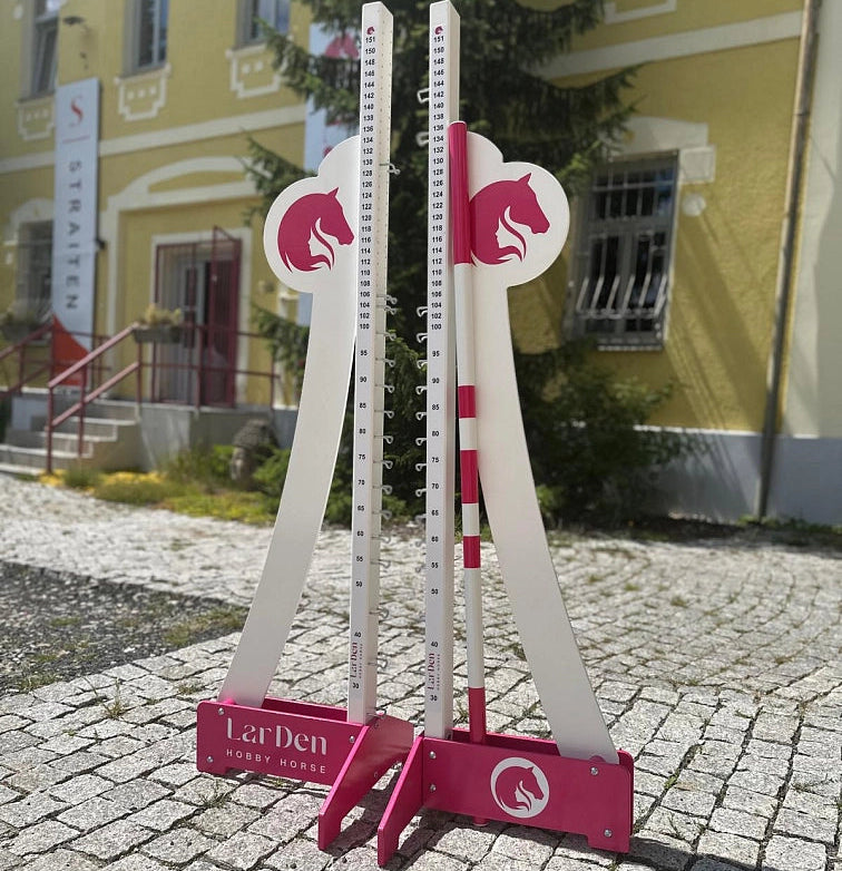 Two white and pink adjustable jump stands for Hobby Horsing are set up on a cobblestone path outdoors, in front of a yellow building with stairs and a sign in the background.