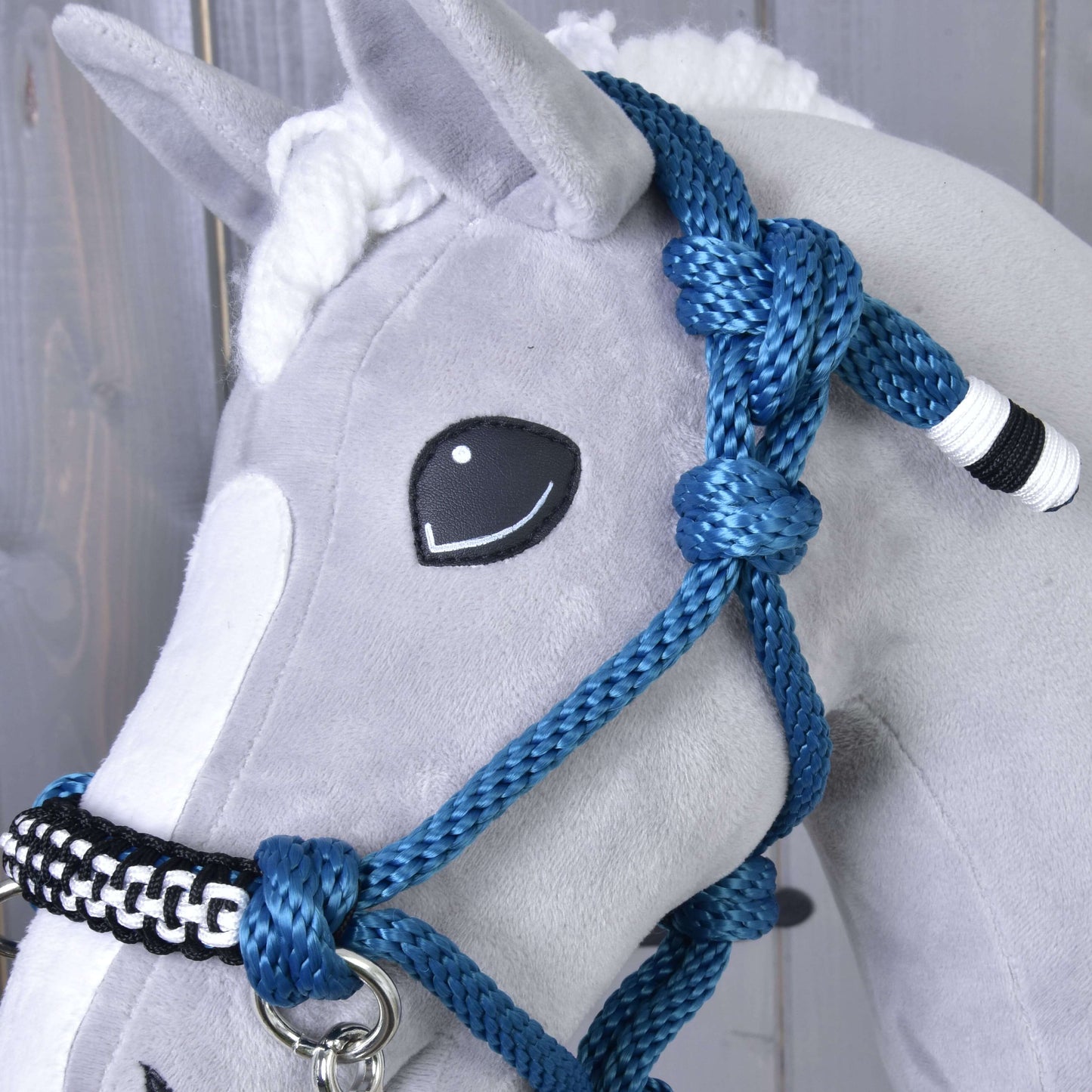 Zbliżenie na szarego pluszowego konia od Hobby Horse LarDen z zestawem Rope set Chess — ręcznie wykonanym kantarem, wodzami, cordeo i niebieską plecioną linką z białymi i czarnymi akcentami. Koń ma wyszywany uśmiech i białą grzywę na tle drewna.