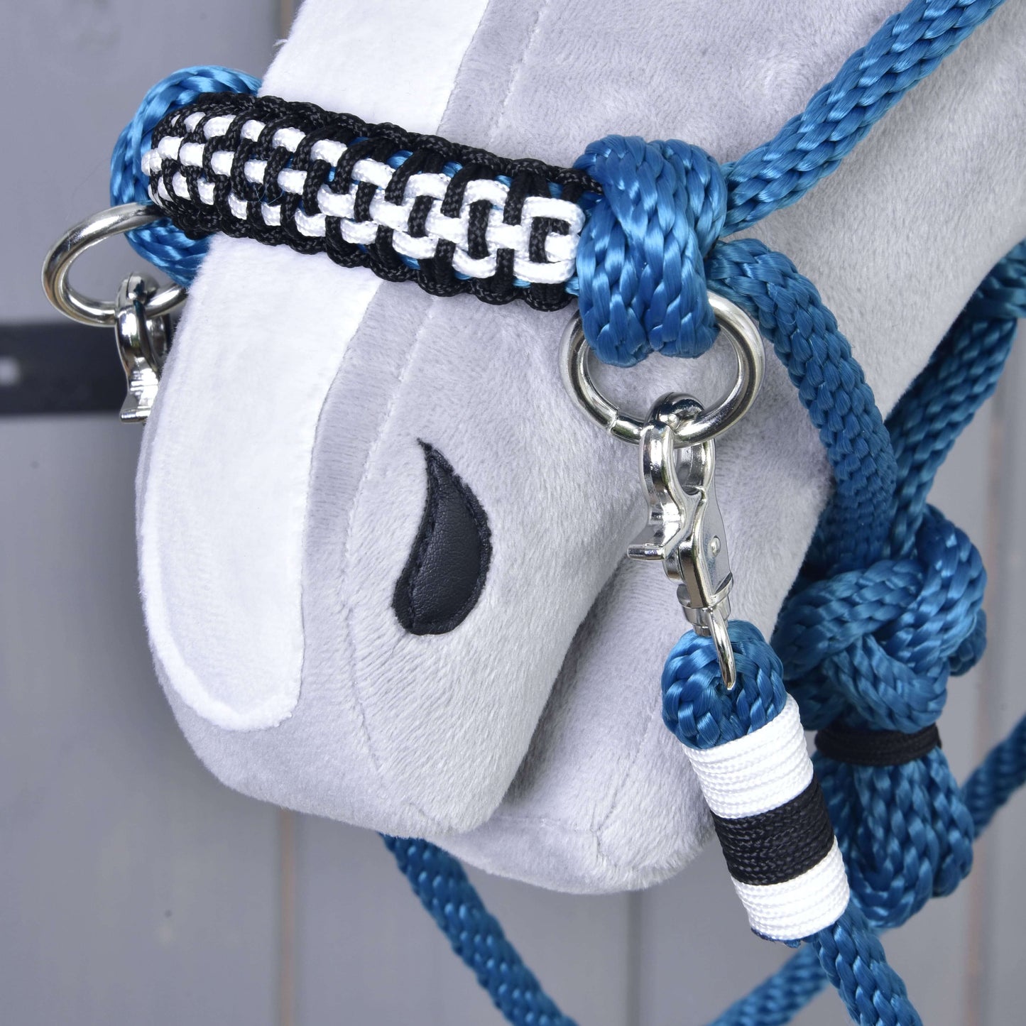 Zbliżenie na miękką szarą głowę konia z wyszywanym czarnym okiem, ubraną w Hobby Horse LarDen Rope set Chess — ręcznie wykonany kantar w kolorach niebieskim, czarnym i białym z metalowymi kółkami i karabińczykami.