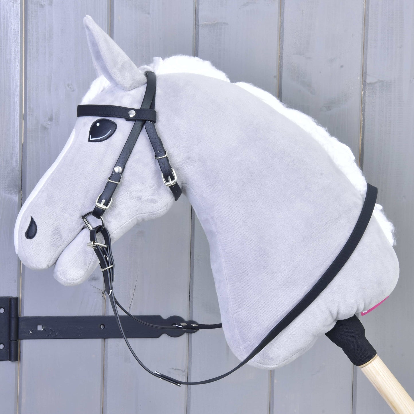 A close-up of a gray hobby horse toy by Hobby Horse LarDen, featuring a Pure Black Complete bridle without noseband and detachable reins, mounted on a wooden stick against a gray wooden background.
