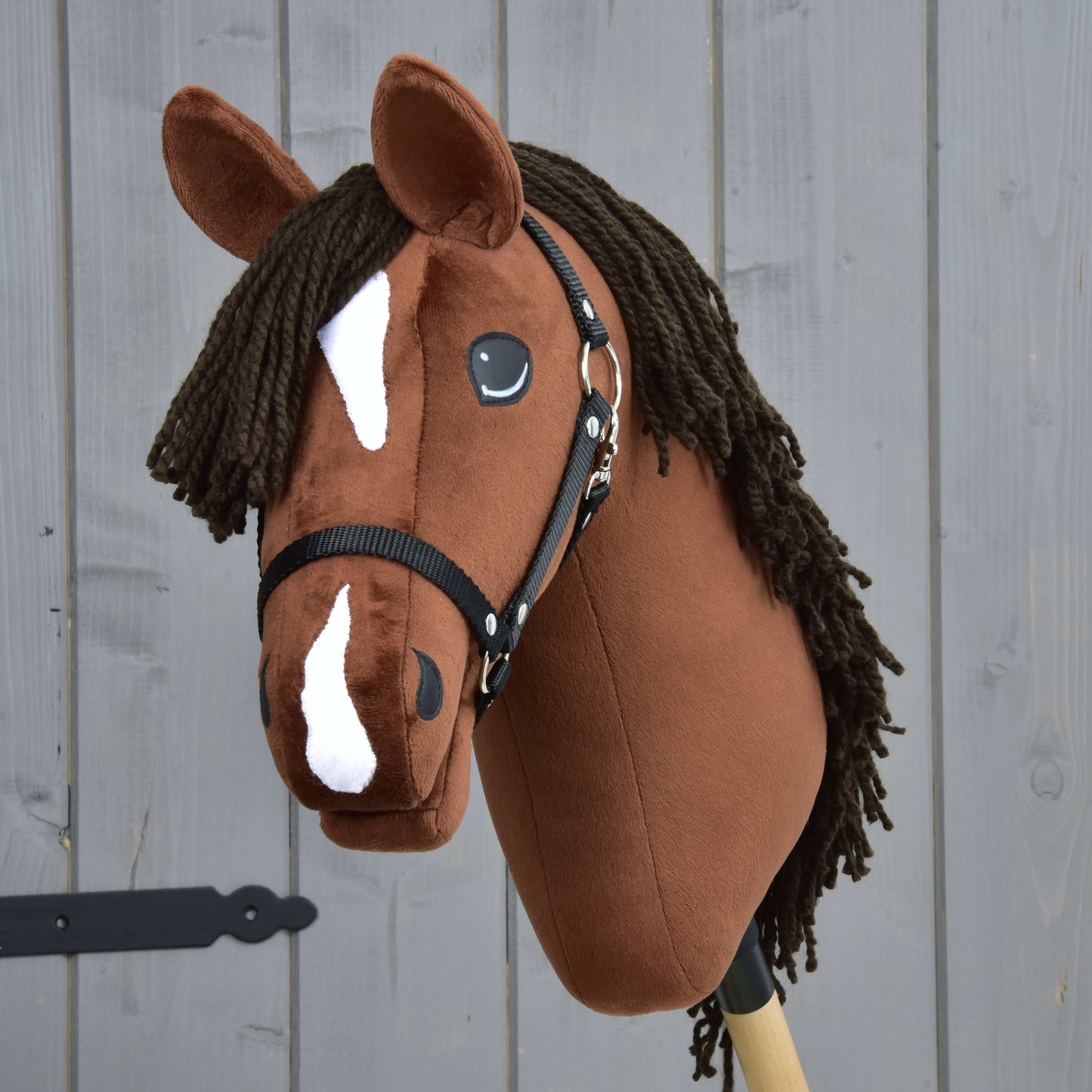 Rocky z Czarnym Kantarem od Hobby Horse LarDen ma brązową głowę w stylu konia pełnej krwi angielskiej, białe odmiany na pysku i ciemną grzywę z włóczki, pokazany na tle szarego drewna.