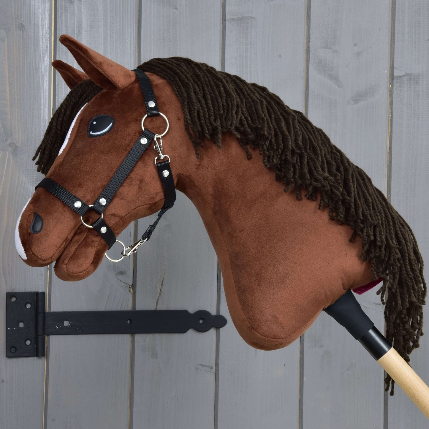 Hobby Horse Rocky z Czarnym Kantarem od Hobby Horse LarDen, brązowy pluszowy konik na kiju, jest pokazany obok szarej drewnianej ściany.
