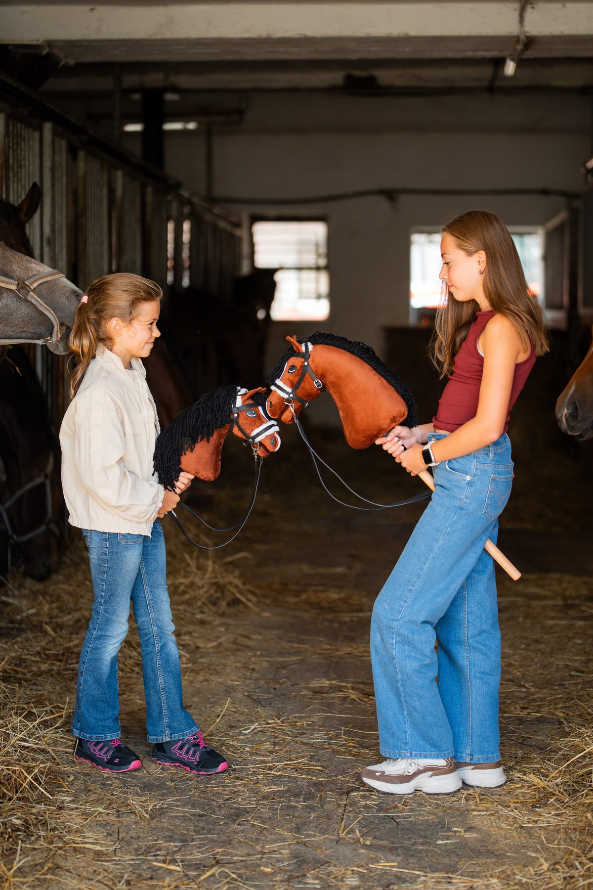 Dwie dziewczynki w korytarzu stajni, każda z kasztanowatym Hobby Horse z czarną grzywą w czarnym ogłowiu z białym obszyciem