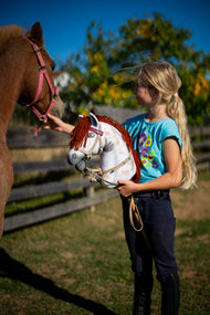 Lifestyle photo: a girl in a turquoise T-shirt holds the Hobby Horse Rufus with Black Halter by Hobby Horse LarDen - white Appaloosa-spotted plush head with a red yarn mane - beside a chestnut pony in a sunny paddock, shown here with the Bordeaux leatherette set - bridle, reins and martingale.