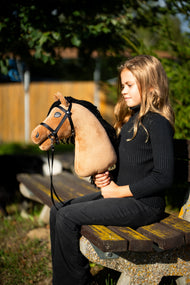 Dziewczyna w czarnym swetrze siedzi na ławce w parku w wieczornym słońcu, trzymając Hobby Horse Samantha z kantarem Black Halter od Hobby Horse LarDen – piaskowa pluszowa głowa, czarna grzywa z włóczki – pokazany z czarnym Ogłowiem z Wodzami paskowymi