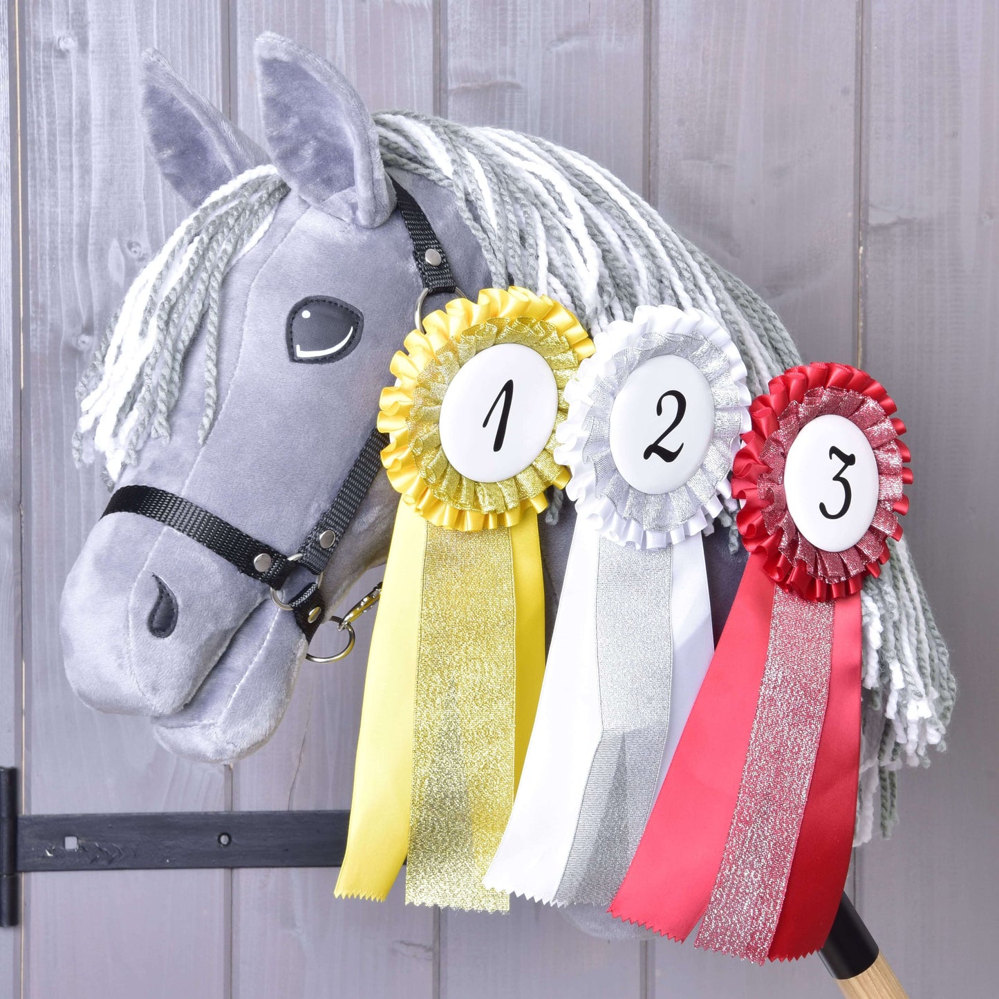 Szary pluszowy Hobby Horse z białą grzywą z włóczki prezentuje Zestaw Wstążek Hobby Horse NOWOŚĆ od Hobby Horse LarDen — żółtą, białą i czerwoną wstążkę za pierwsze, drugie i trzecie miejsce — zawieszone na szyi na tle drewna.