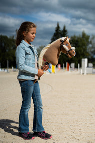 Dziewczyna w dżinsowej kurtce stoi na piaszczystym placu, trzymając Hobby Horse Smart z kantarem Black Halter od Hobby Horse LarDen, a za nią leżą drążki do skoków – głowa z pluszu palomino, długa kremowa grzywa z włóczki – pokazany z czarnym kantarem ze skóry ekologicznej z pełnym futerkiem