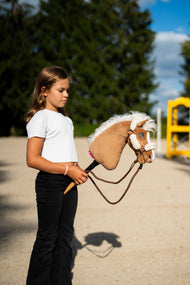 Dziewczyna w białej bluzce prowadzi Hobby Horse Star z czarnym kantarem od Hobby Horse LarDen przez piaszczysty plac, a za nią stoi żółty stojak do przeszkody – głowa z pluszu palomino, falująca biała grzywa z włóczki – pokazany z brązowym kantarem z pełnym futerkiem i Wodzami Vivaldi