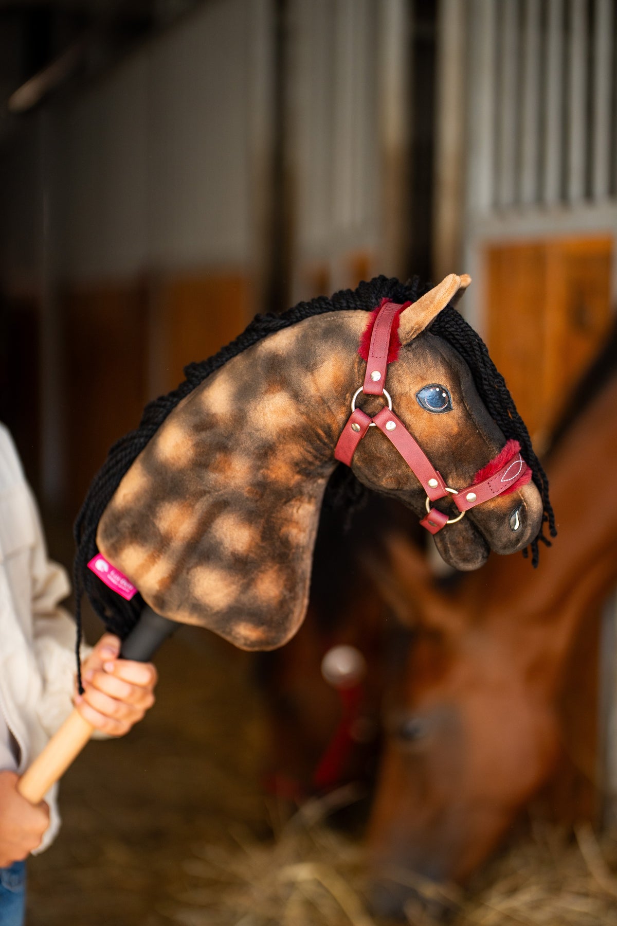 Głowa Hobby Horse Stormy z kantarem Black Halter od Hobby Horse LarDen, uniesiona w stajni, z rozmytym prawdziwym gniadym koniem w tle – dereszowaty brązowy plusz, niebieskie oko, czarna grzywa z włóczki – pokazany z kantarem z futerkiem Burgundy ze skóry ekologicznej